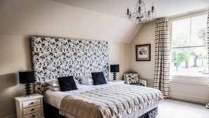 Un dormitorio con una cama grande, dos mesitas de noche, una araña, una ventana con cortinas y un cabecero estampado con motivos florales.