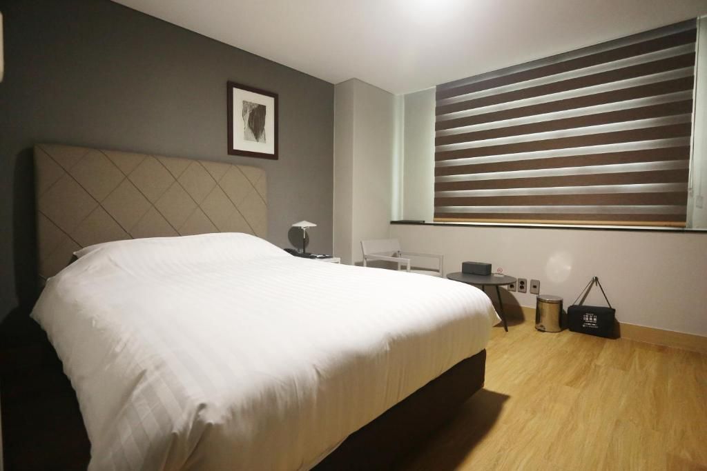 A hotel room with a bed, bedside table, lamp, window with blinds, and a small trash bin.