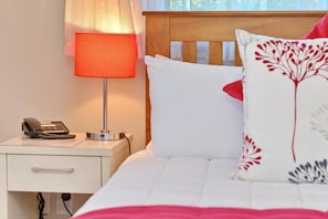 A neatly made bed with white bedding and a decorative pillow, a bedside lamp, a telephone, and a drawer unit.