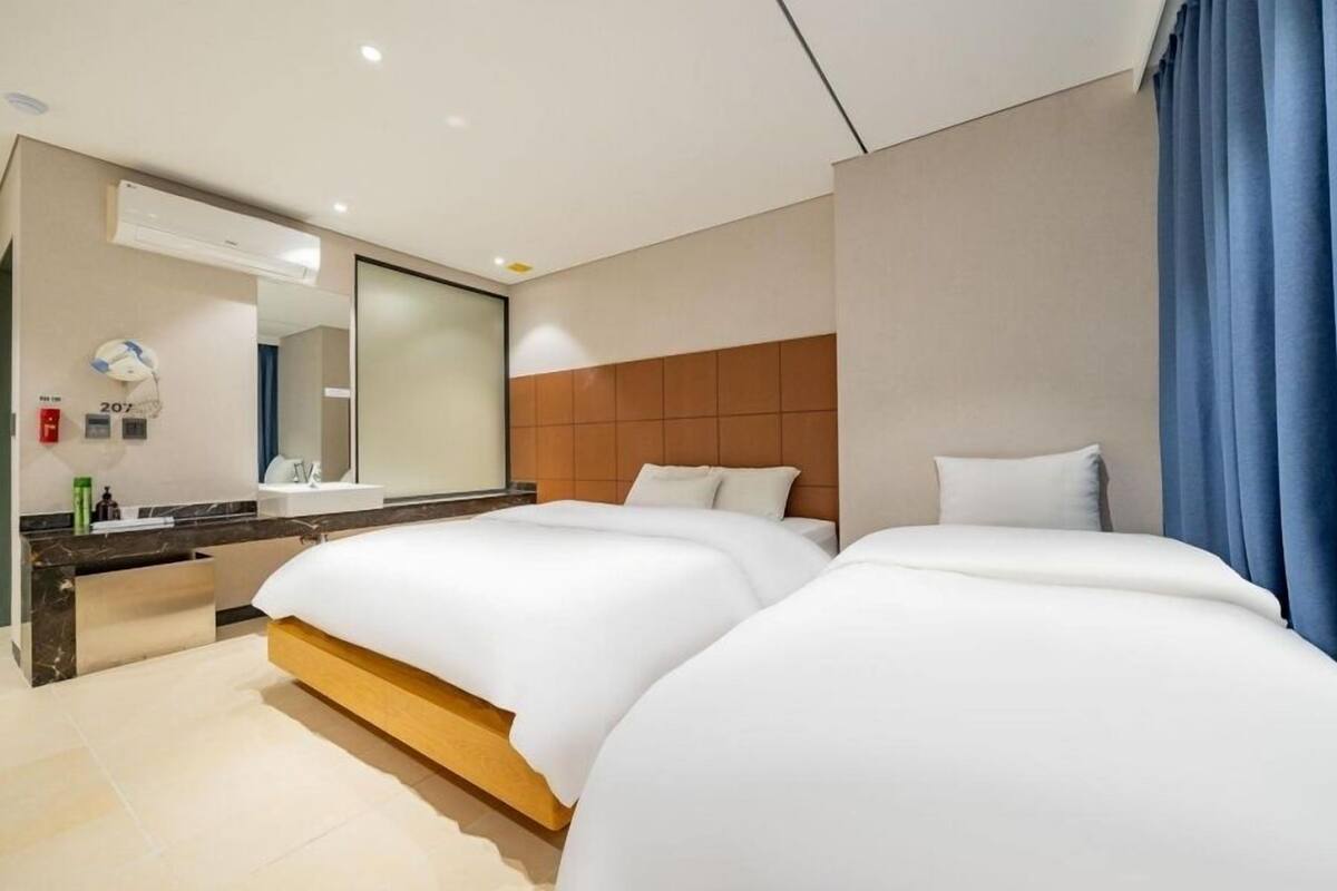 Une chambre d’hôtel avec deux lits, un grand miroir, une tête de lit en bois, une salle de bain avec un lavabo et un rideau bleu.