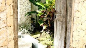 A rustic stone wall with a wooden door leading to a garden area with plants and a concrete pathway. - Mayana Villas (Canggu)