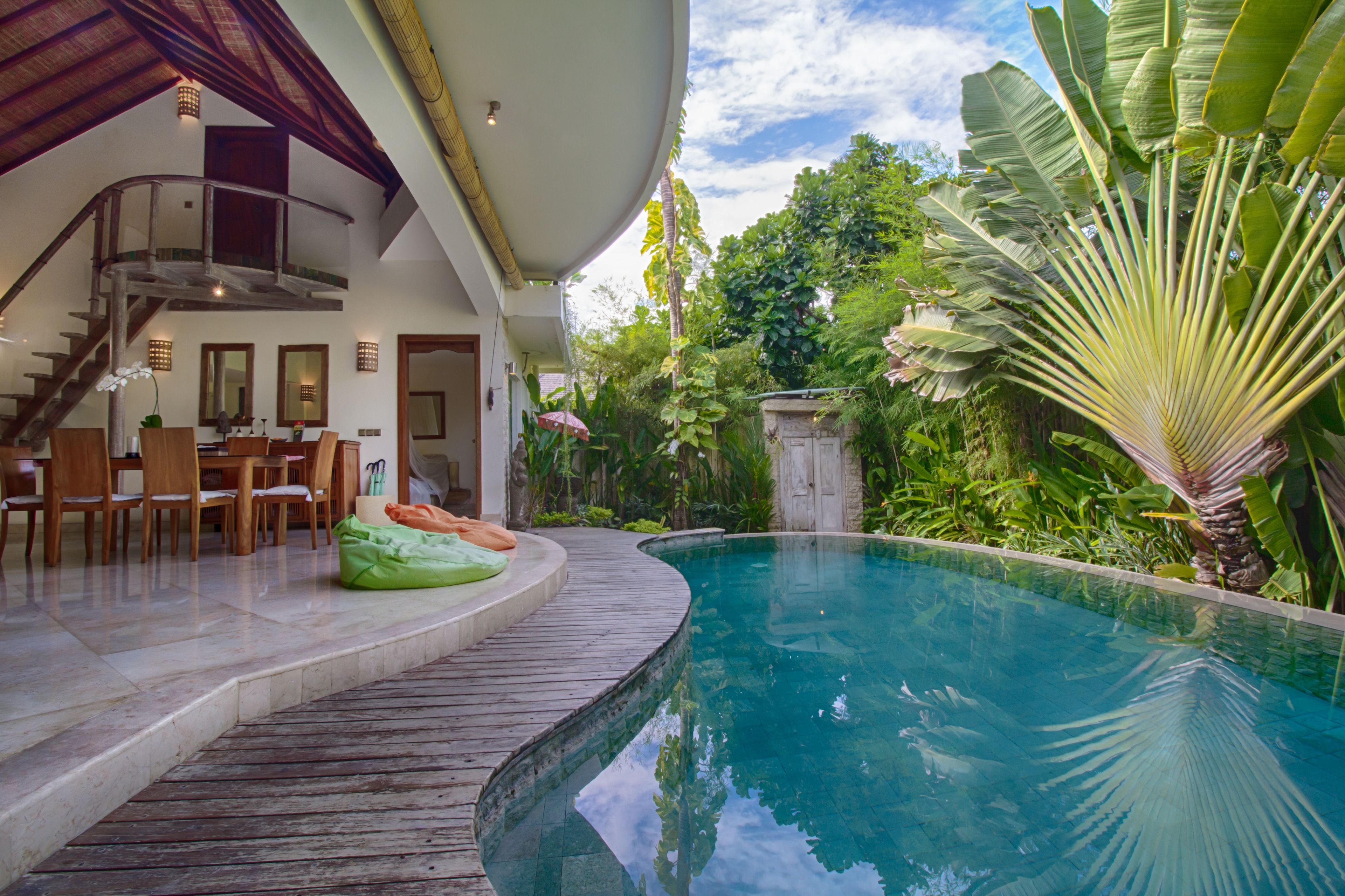 A pool area with a wooden deck, a dining area with chairs, and lush greenery.