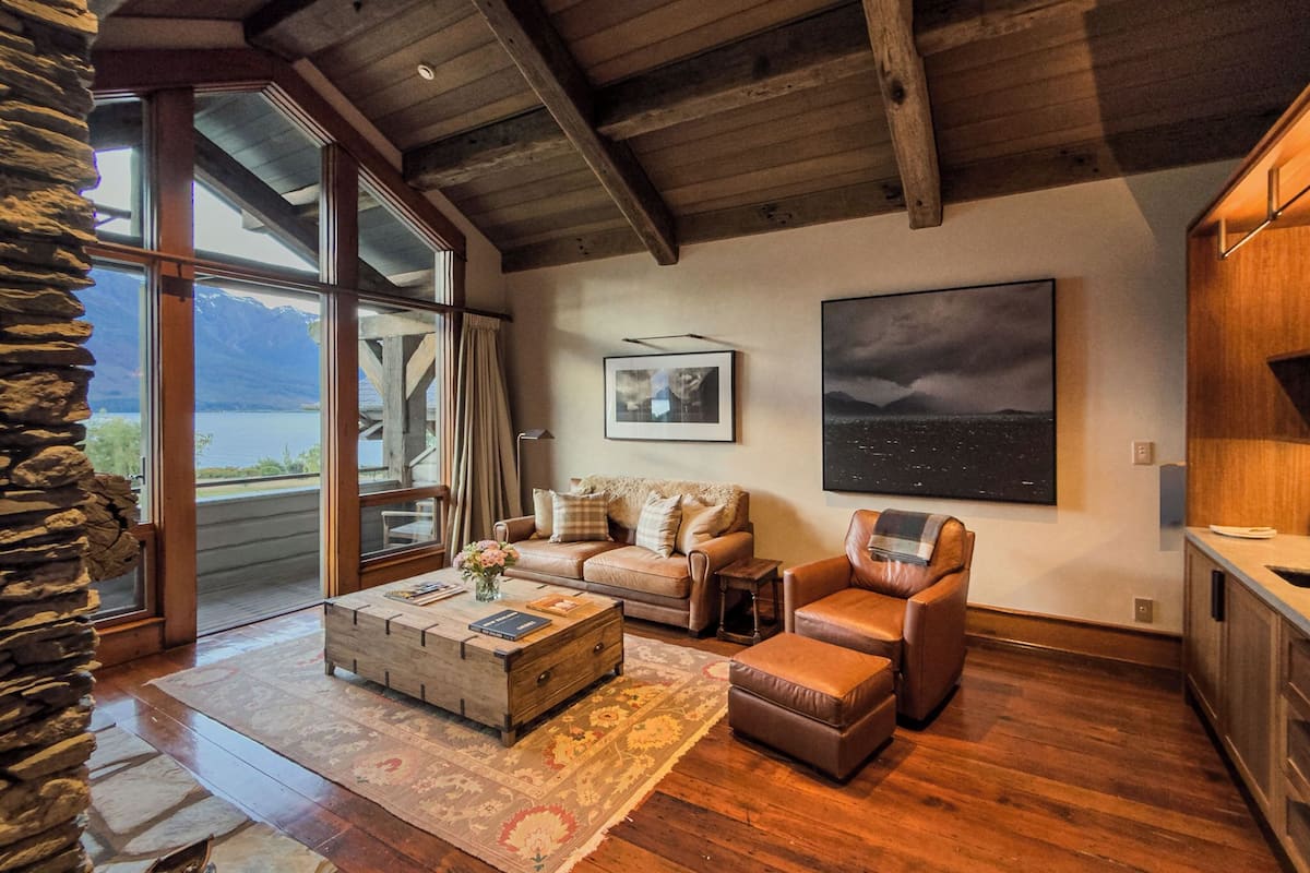 A spacious living area with a stone wall, wooden ceiling beams, a large window with a view of mountains, a sofa, a coffee table, and a fireplace.