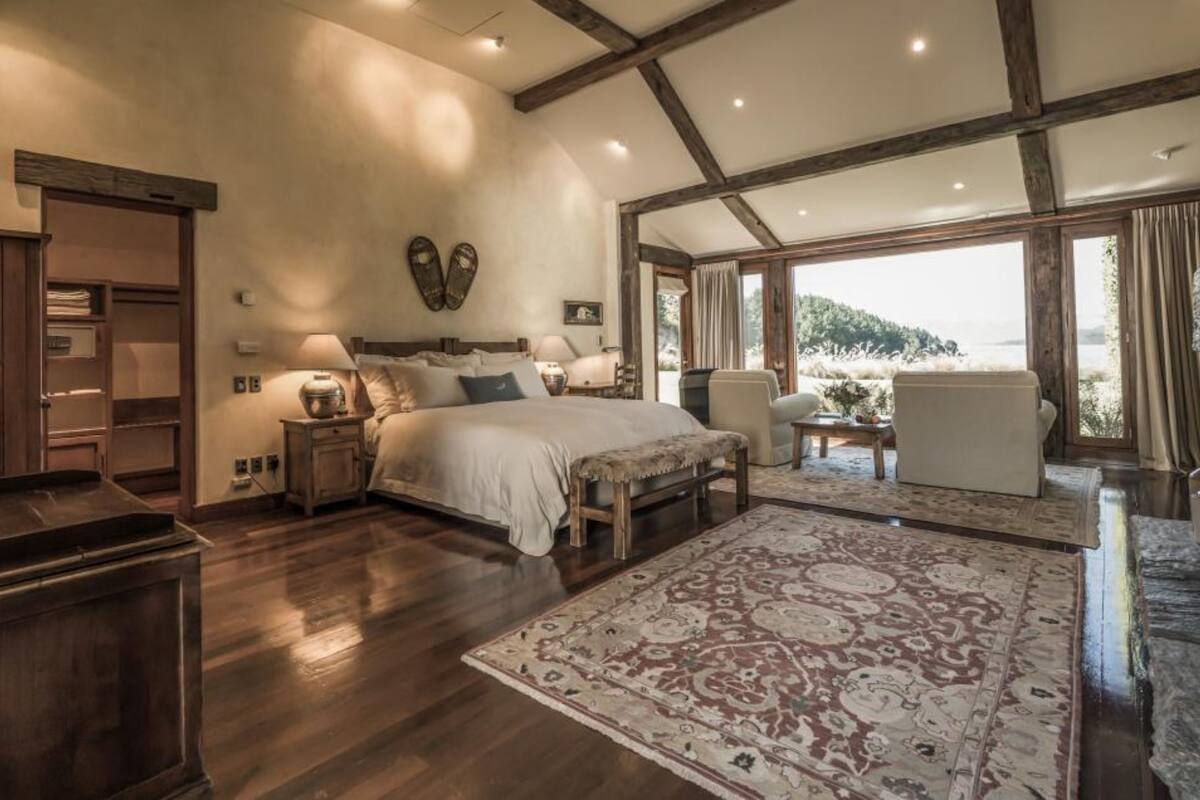 A spacious bedroom with a large bed, a wooden bench, a nightstand, and a desk. There is a large window with a view of the outdoors.