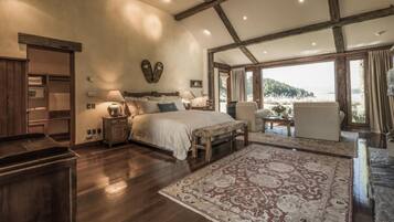 Um quarto espaçoso com uma cama grande, um banquinho de madeira, uma mesa de cabeceira e uma escrivaninha. Há uma janela grande com vista para o exterior.