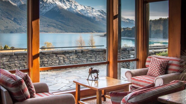 A room with large windows offering a view of a lake and mountains. There is a wooden table with a decorative item on it, and striped upholstered furniture. - Blanket Bay (Glenorchy)