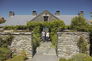 Traditional stone building with a steeply pitched roof and two chimneys, set amidst lush greenery and flowering plants.