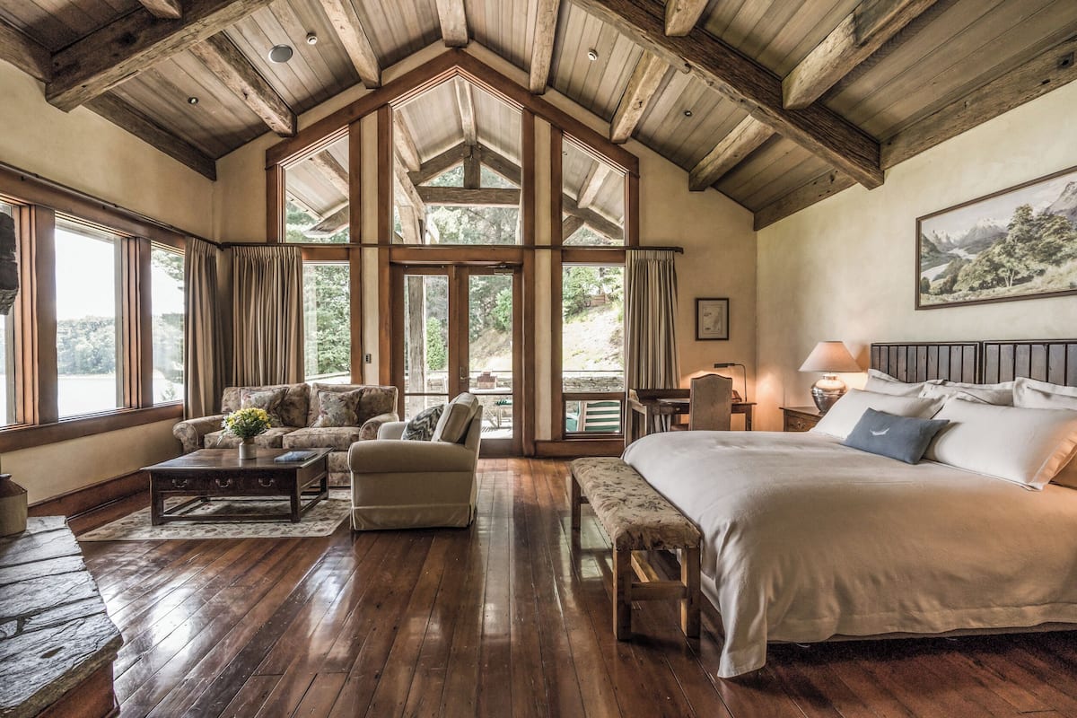 A spacious room with a large bed, a sofa, a coffee table, and a bench. There are large windows and a wooden ceiling.