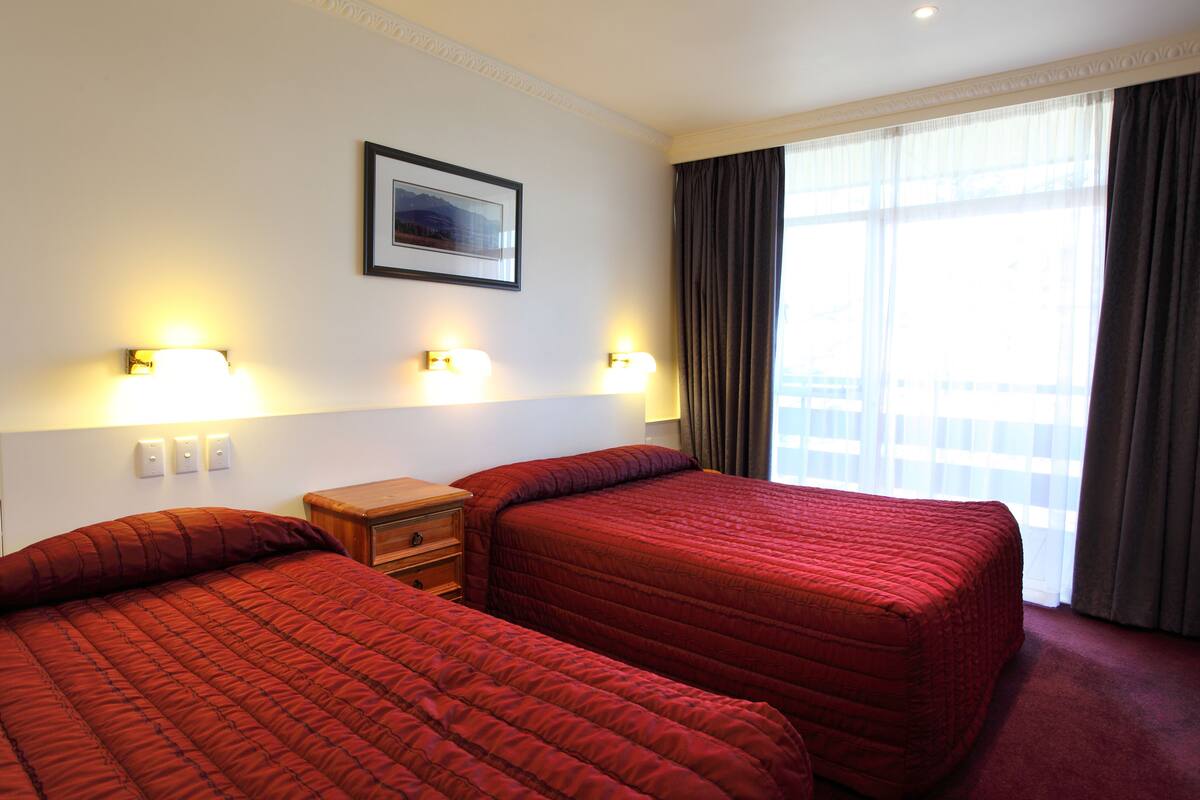 A hotel room with two beds, a nightstand, wall-mounted lights, a framed picture, and a large window with curtains.