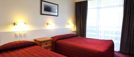 A hotel room with two beds, a nightstand, wall-mounted lights, a framed picture, and a large window with curtains.