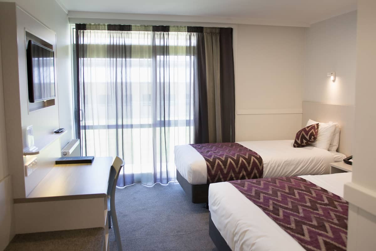 A hotel room with two beds, a desk, a chair, a TV, and a window with curtains.
