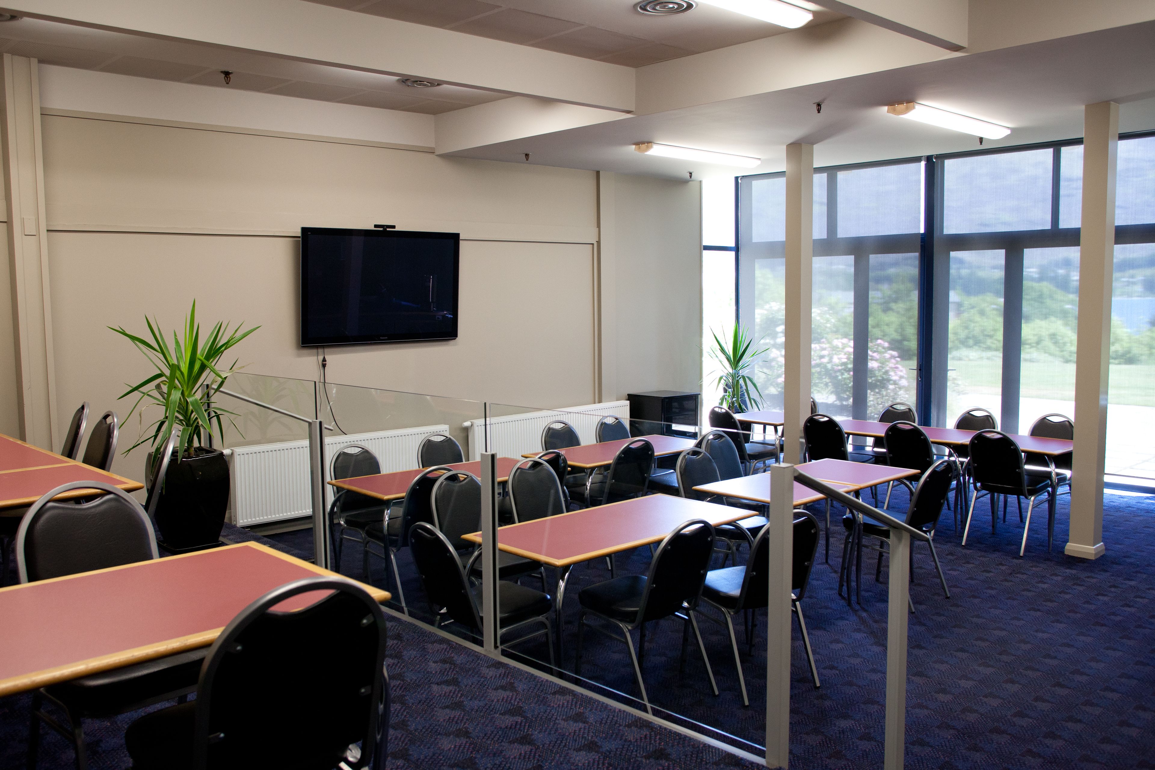 A conference room with tables and chairs, a large screen, and large windows with a view of greenery.