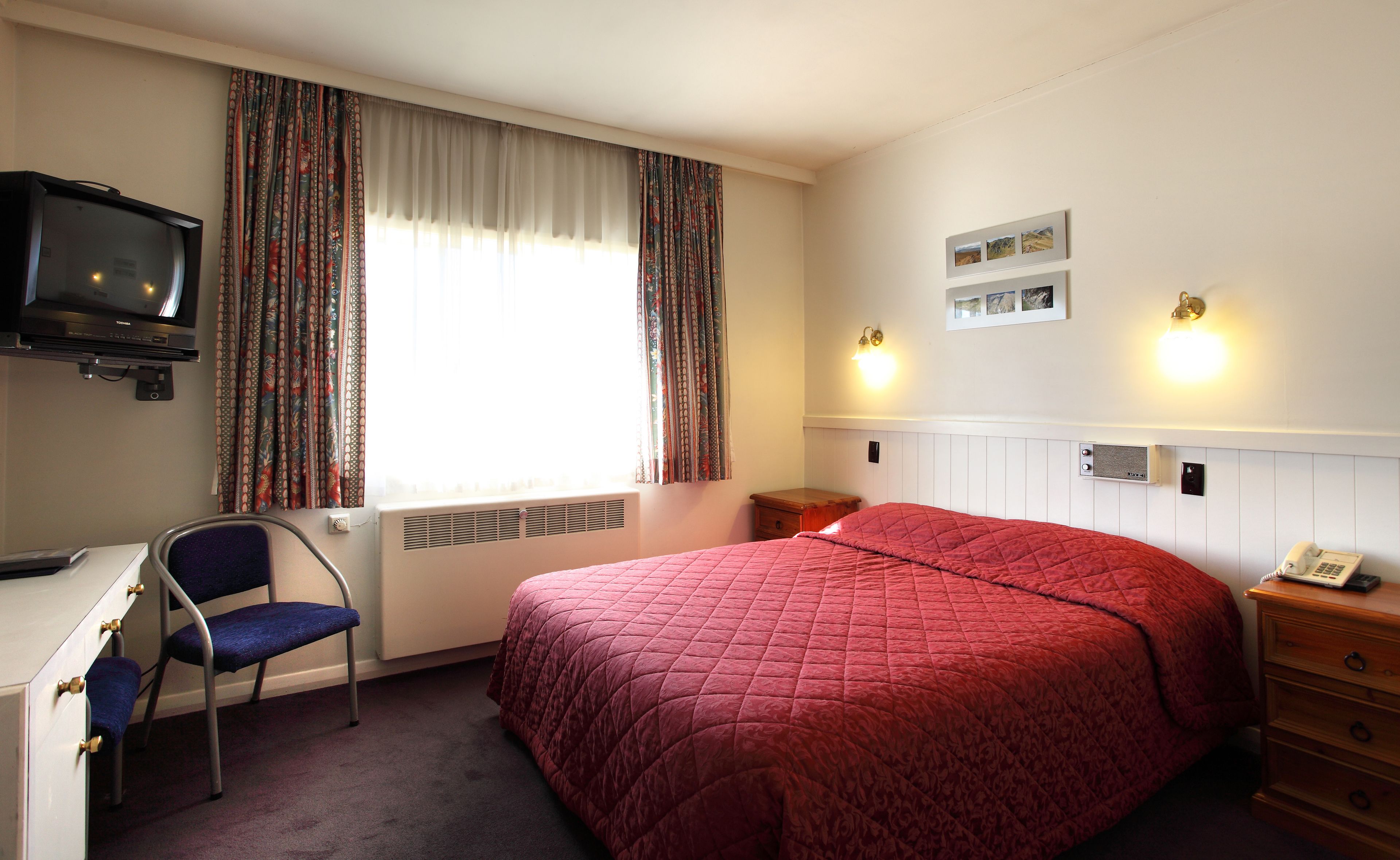 A hotel room with a bed, a desk with a chair, a television, and a window with curtains.