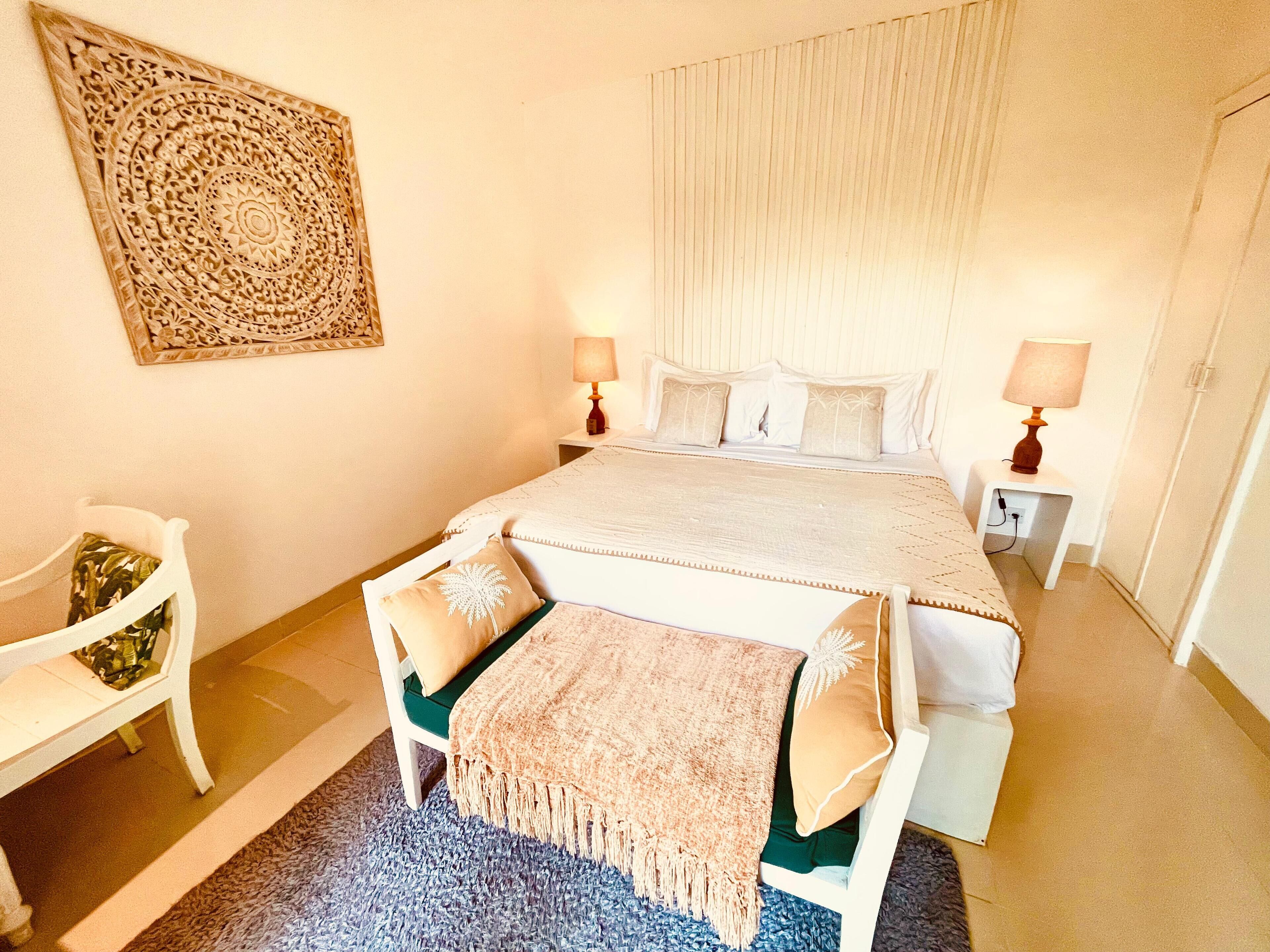 Un dormitorio con cama, dos mesitas de noche con lámparas, una silla, un adorno en la pared y una alfombra azul.