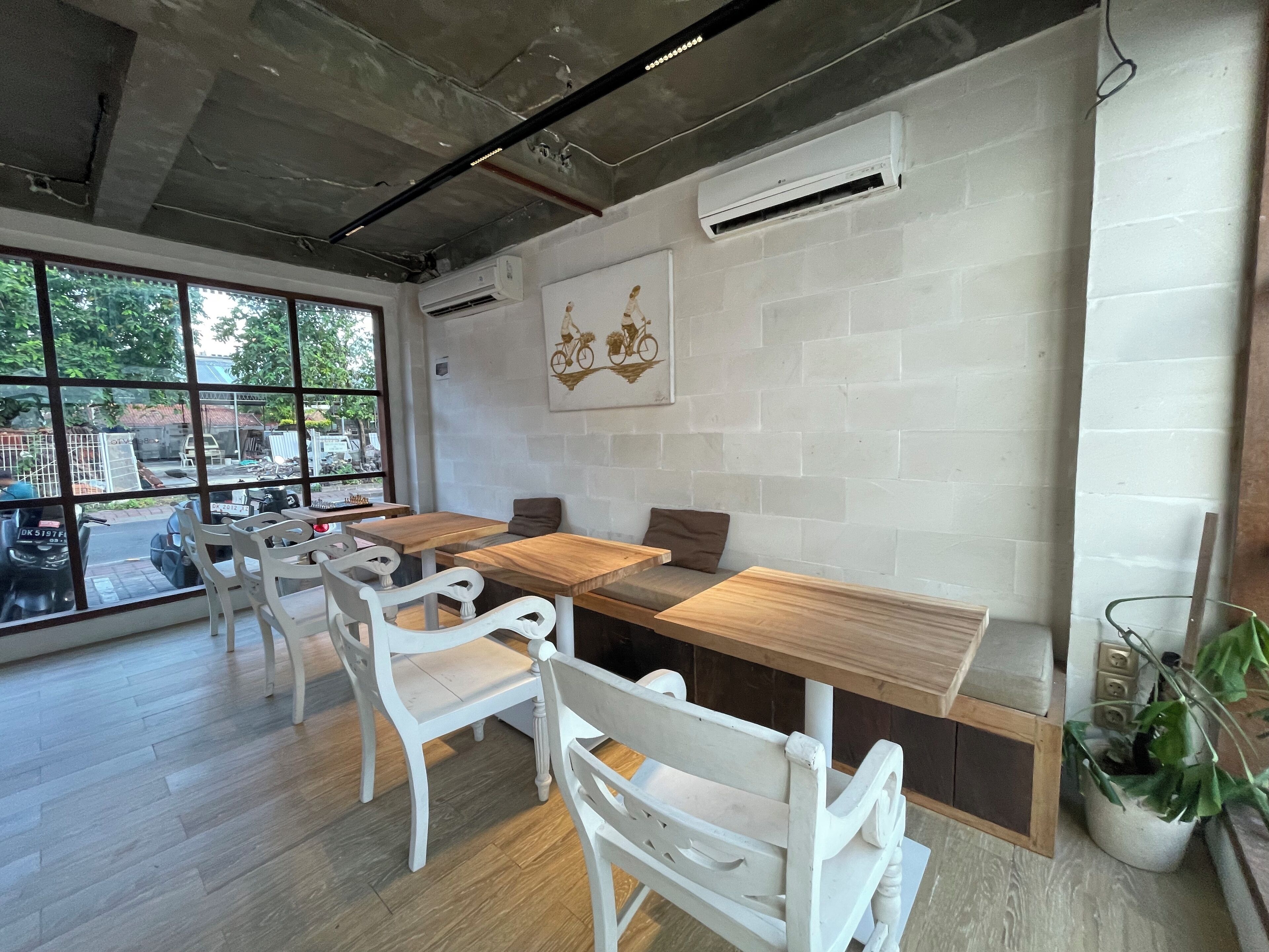 Una cafetería con mesas de madera y sillas blancas, ventanas amplias y una pared de ladrillo con un mural de una bicicleta.