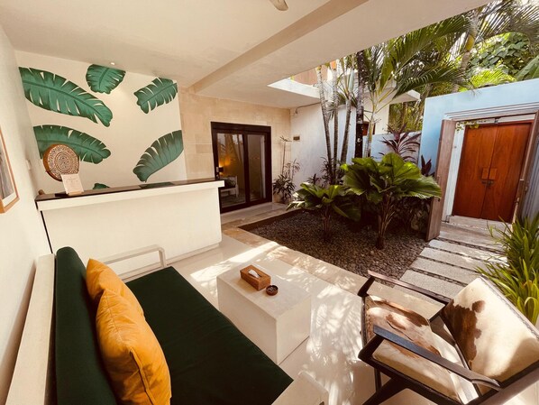 A modern interior with a reception desk, a seating area with a sofa and armchair, and a garden with tropical plants.