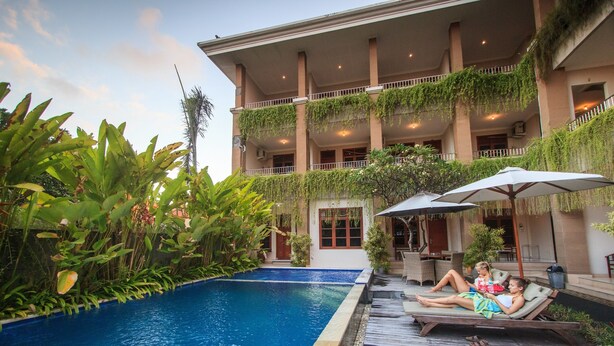 Zona de piscina con sillones de descanso y sombrillas, rodeada de exuberante vegetación y un edificio de varios pisos con balcones.