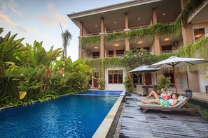 A pool area with lounge chairs and umbrellas, surrounded by lush greenery and a multi-story building with balconies. - Pondok Anyar Inn (Kuta)