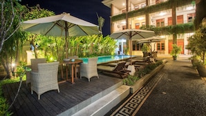 Zona de piscina exterior con mobiliario de mimbre y sombrillas, rodeada de abundante vegetación y un edificio bien iluminado al fondo.
