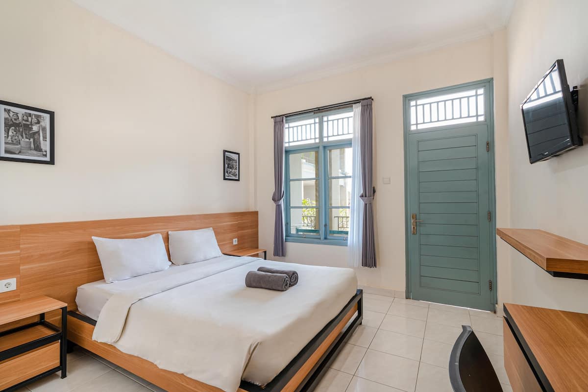 A neatly arranged bedroom with a bed, bedside tables, a window with curtains, a door, and a mounted TV.