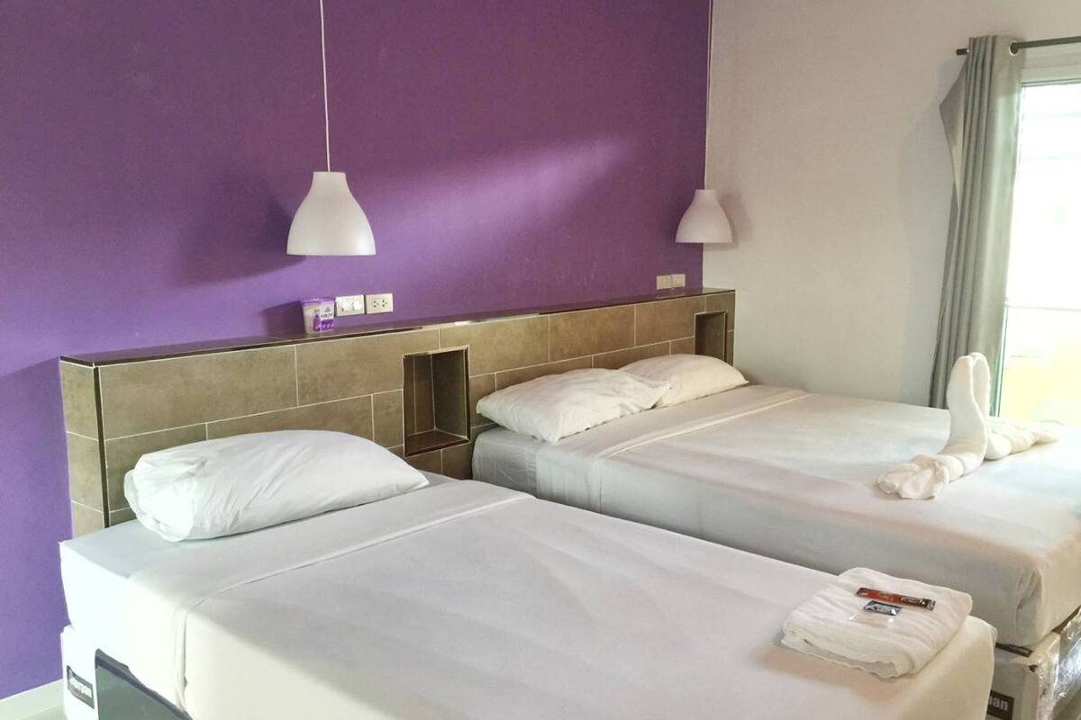 Une chambre d’hôtel avec deux lits, un mur violet et une fenêtre avec des rideaux.