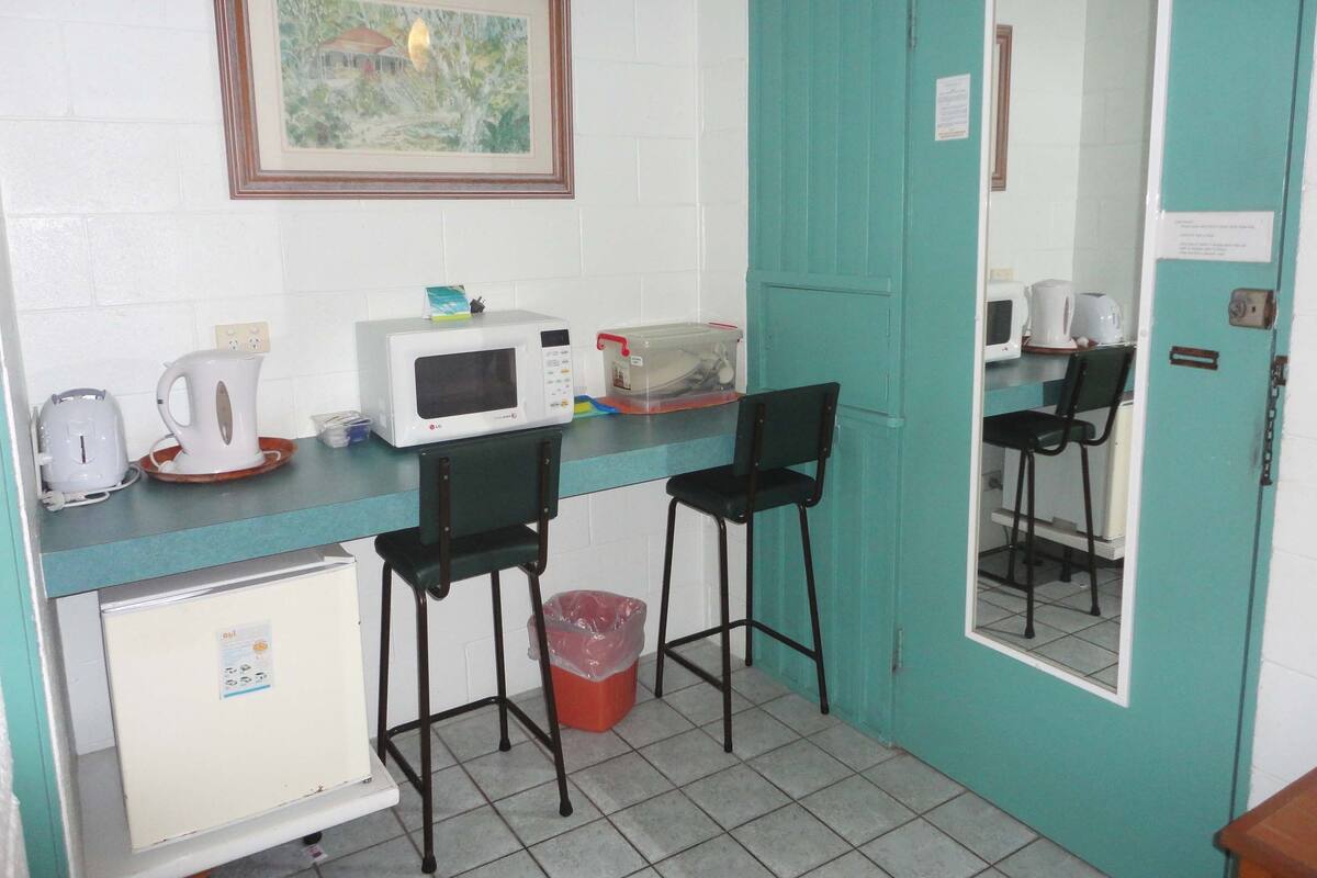A compact kitchenette with a microwave, kettle, and toaster on the counter, two chairs, a small refrigerator, and a trash bin.