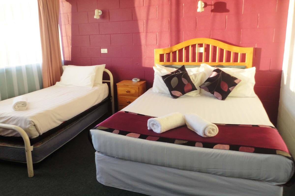 A room with two beds, one with a wooden headboard and the other with a metal frame. There is a nightstand with a clock between the beds.