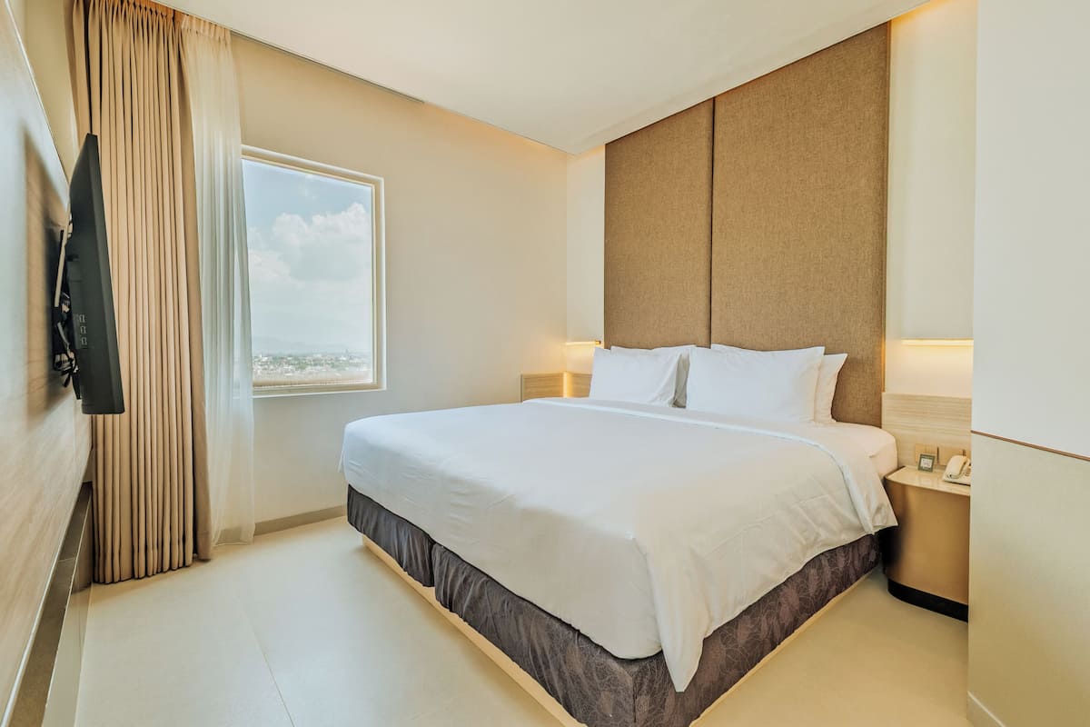 Una habitación de hotel con una cama grande, una mesita de noche, una lámpara, una ventana con cortinas y vistas a la ciudad.