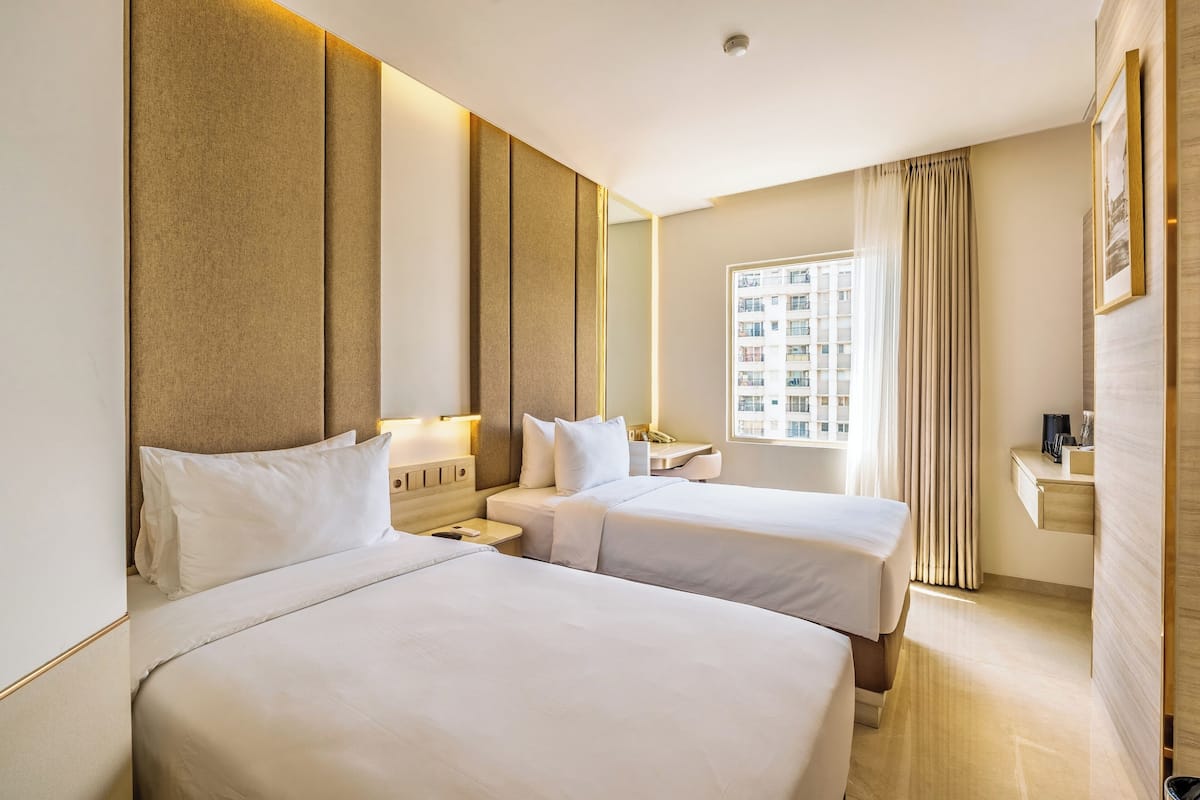 Habitación de hotel con dos camas, una mesita de noche, un escritorio, una silla, una ventana con cortinas y un televisor montado en la pared.