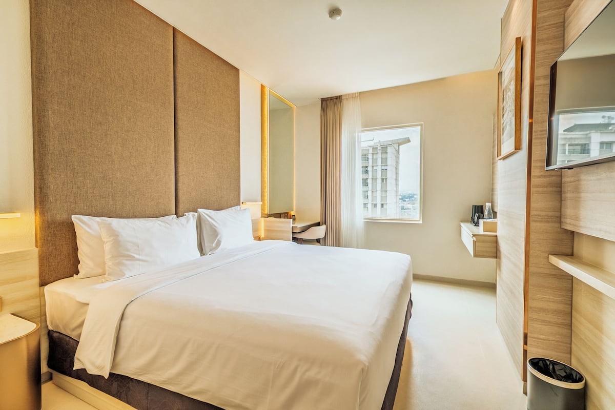 Habitación de hotel con una cama grande, un escritorio, un espejo, un televisor y una ventana con vistas a la ciudad.