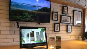 Un mostrador de recepción de hotel con un monitor de computadora que muestra un recorrido virtual, un televisor arriba que muestra una vista panorámica del océano y cuadros enmarcados en la pared.