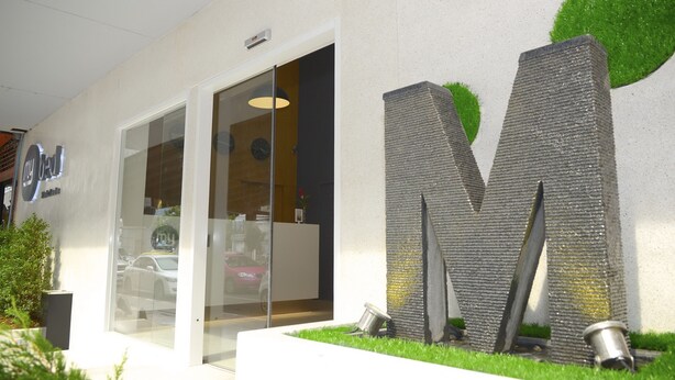 Entrada de un edificio moderno con puerta de vidrio, una escultura grande con la letra 'M' y un letrero que dice "my hotel".