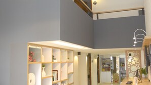 Un interior espacioso y bien iluminado, con una amplia estantería de madera en el centro, una zona de comedor con taburetes y una escalera que sube a un nivel superior.