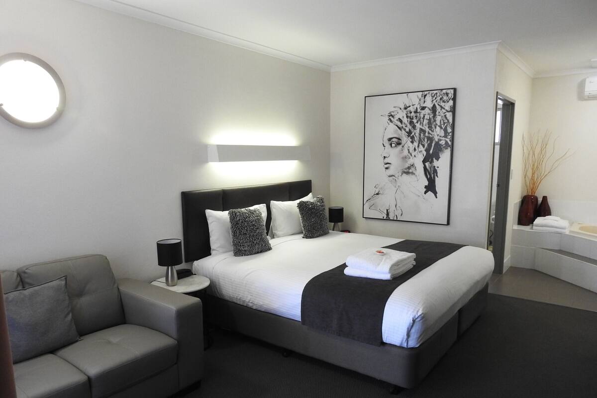 A modern hotel room with a large bed, a bedside table with a lamp, a grey sofa, and a large abstract painting on the wall.