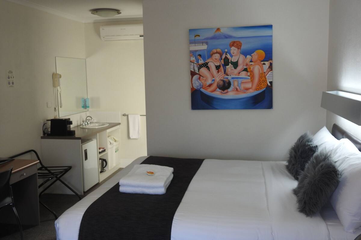 A hotel room with a bed, a desk, a chair, a sink, a mirror, and a painting on the wall.