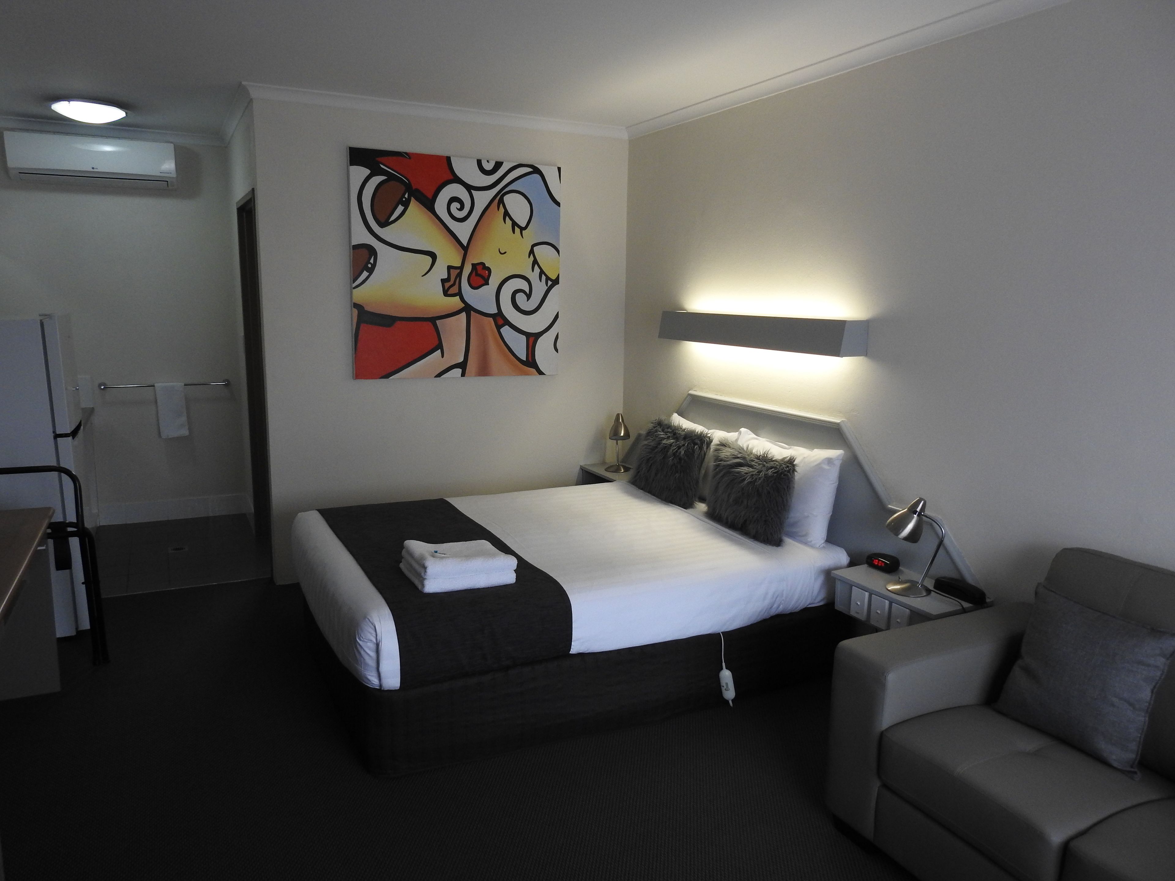 Una habitación de hotel con cama, mesita de noche con lámpara, sofá y un cuadro en la pared.