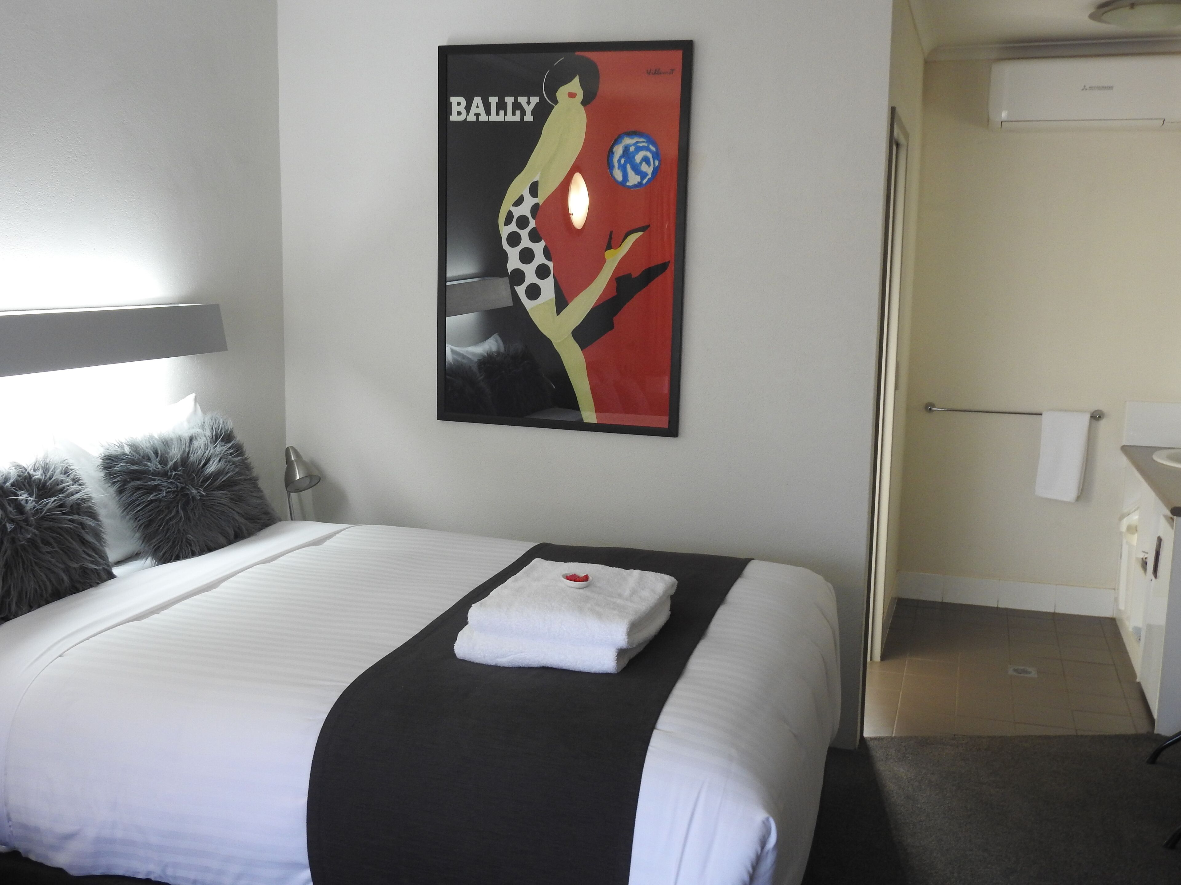 Una habitación de hotel con una cama bien hecha, una obra de arte enmarcada en la pared y un baño a la vista a través de una puerta abierta.