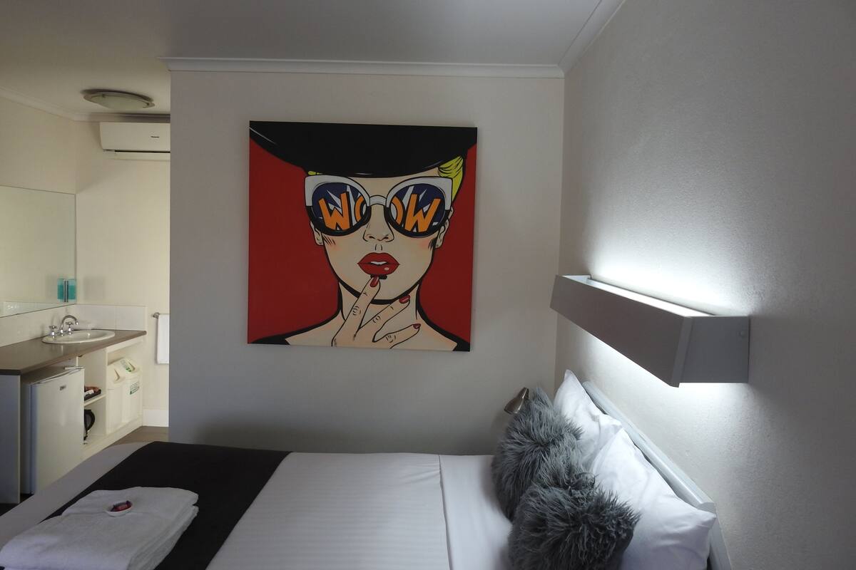 A hotel room with a bed, a painting on the wall, a bathroom with a sink and mirror, and a white ceiling.