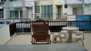 A balcony with a wooden chair and small stools, a table, and a view of a multi-story building with balconies and windows. - Hill Inn (Pattaya)