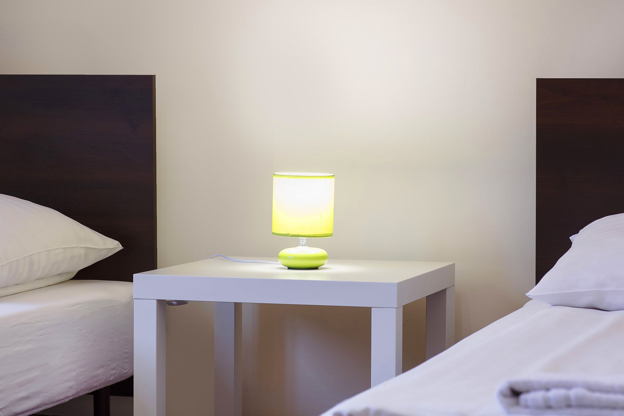 Una cama bien tendida con ropa blanca, una mesita de noche con una lámpara verde y una pared lisa.