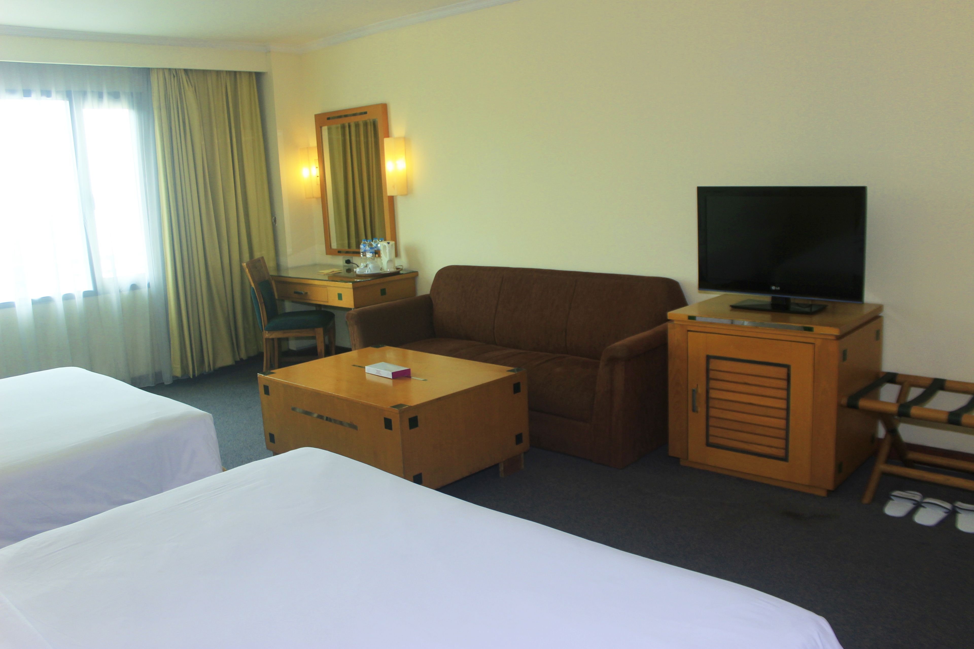 A hotel room with two beds, a wooden desk, a television, a mirror, and a window with curtains.