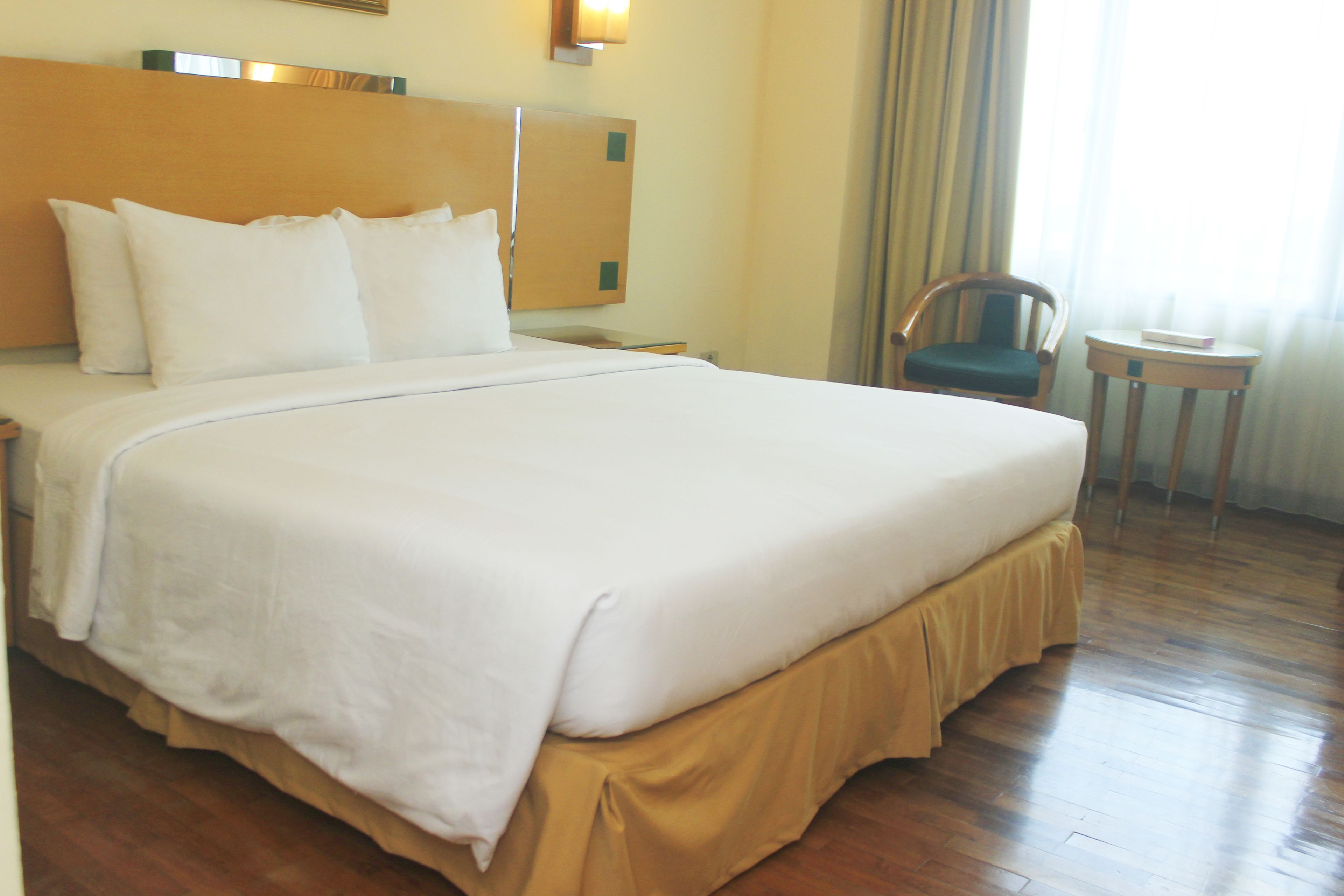 A hotel room with a large bed, a wooden headboard, a nightstand, a chair, a small table, and a window with curtains.