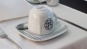 Uma chávena de cerâmica branca com um desenho floral sobre um prato branco, acompanhada por um garfo e uma colher sobre uma toalha de mesa.