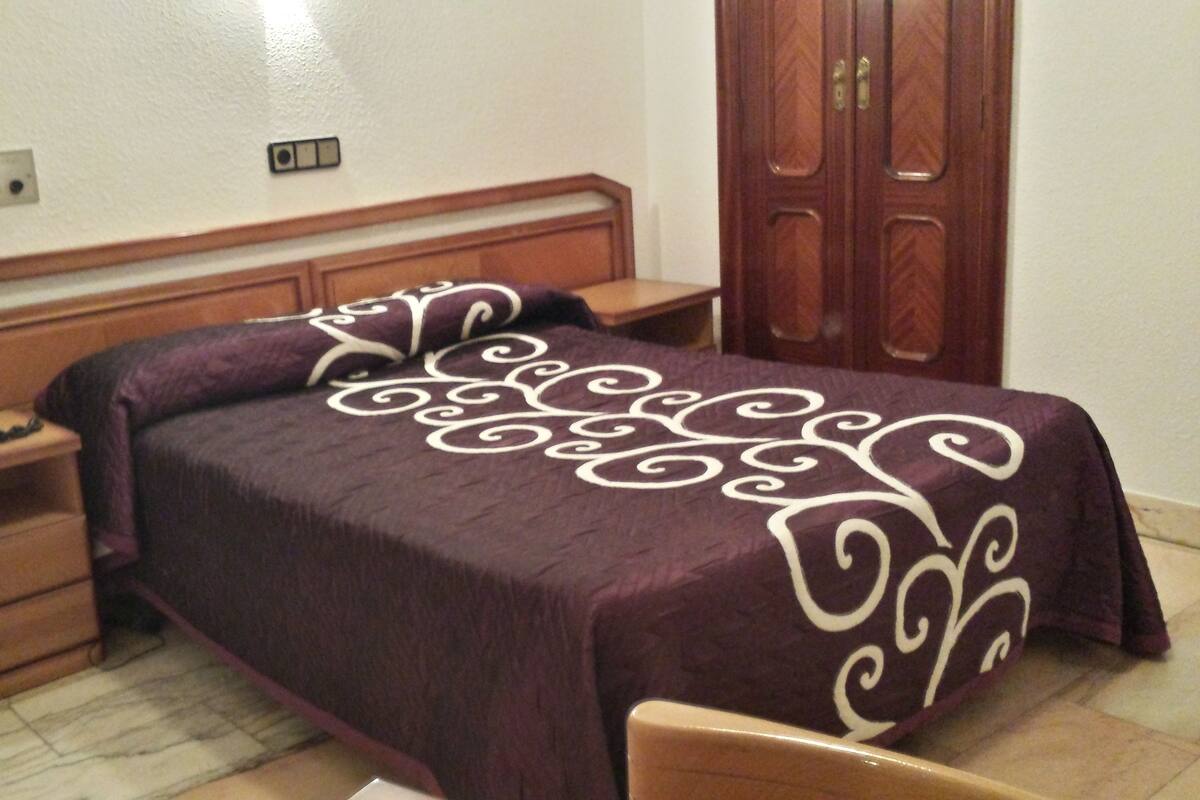 A bedroom with a bed, wooden headboard, a nightstand, a desk, and a wardrobe.