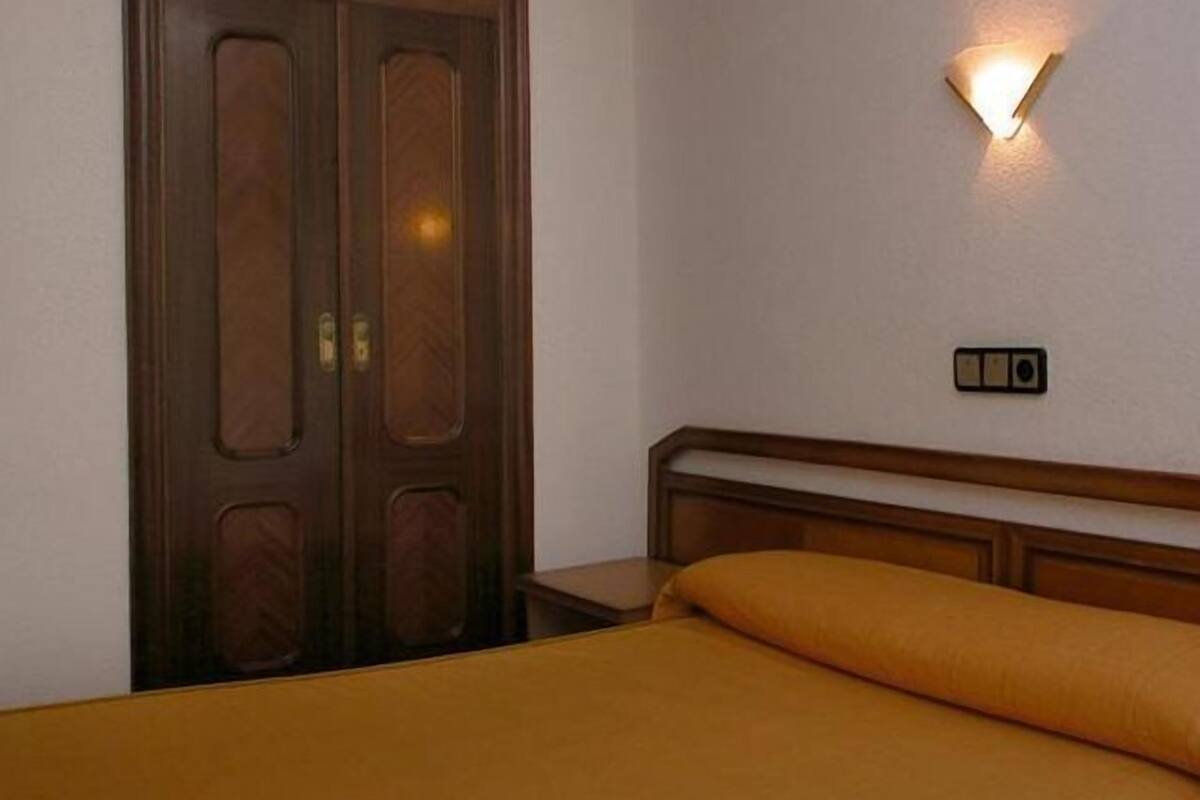 A hotel room with a wooden door, a bed with a yellow bedspread, and a wall-mounted light fixture.