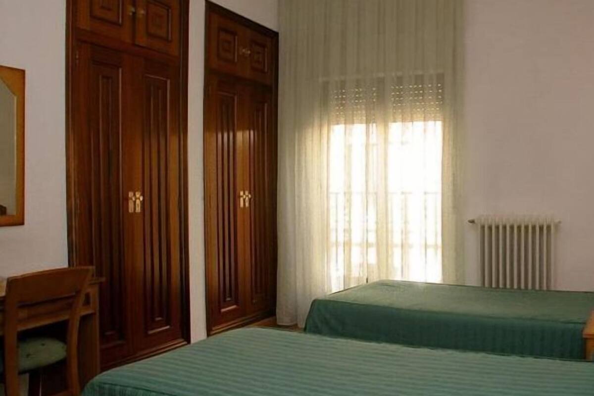 A room with two beds, a wooden wardrobe, a chair, and a window with sheer curtains.