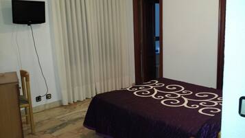 A bedroom with a bed, a wooden chair, a television mounted on the wall, and a window with blinds.
