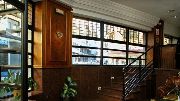 A lobby with a staircase, a reception desk, and a view of the street through large windows.