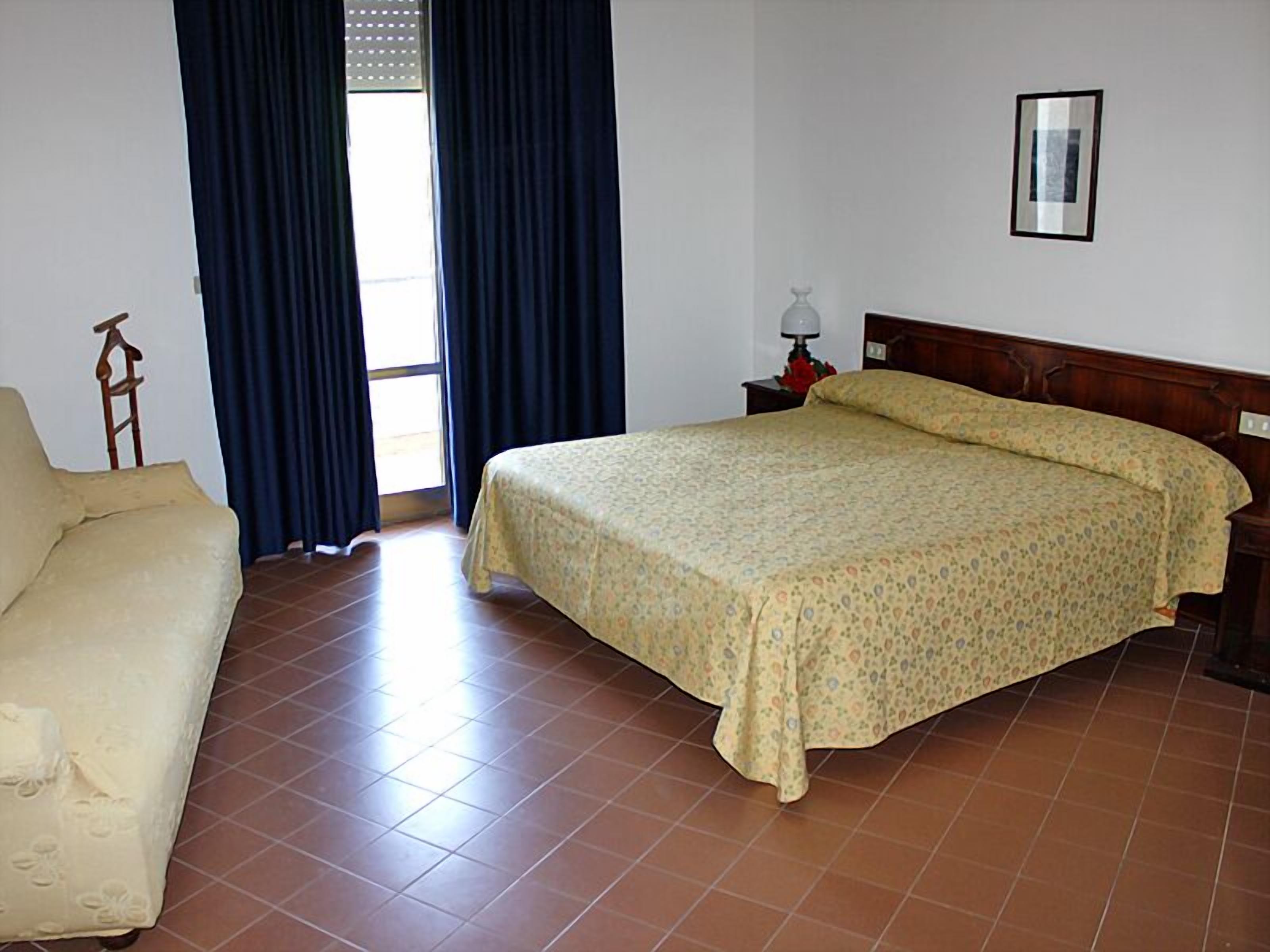 A bedroom with a bed, a chair, a nightstand, a lamp, and a window with curtains.