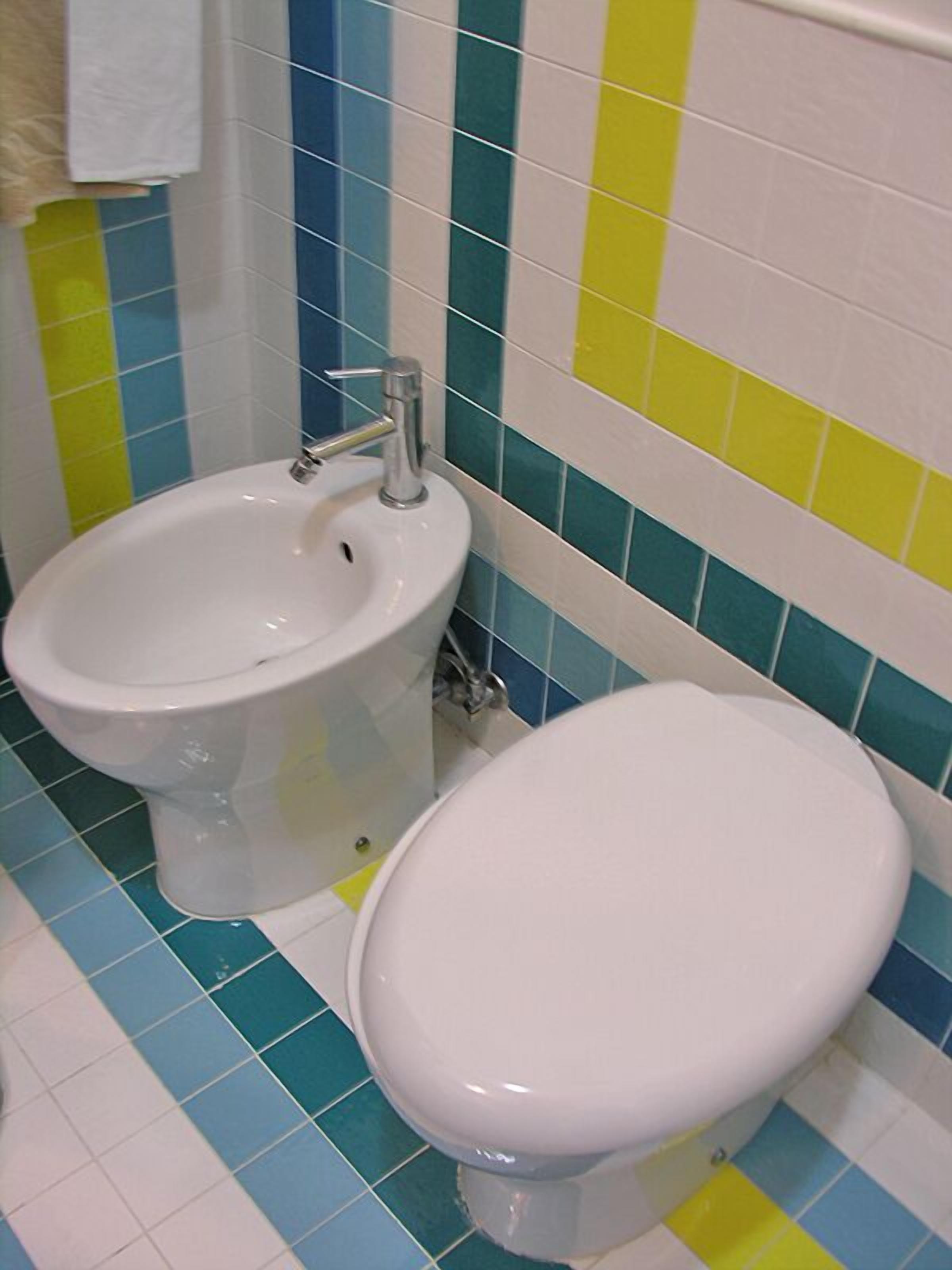 Un baño con bidé, inodoro y paredes cubiertas de azulejos en azul, amarillo y blanco.